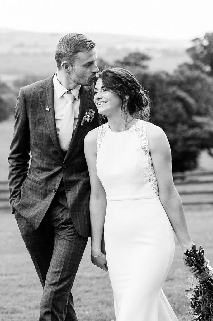 Bride in a jewel neck crepe gown with a fit and flare skirt and lace detail, walks alongside her groom in an open field.