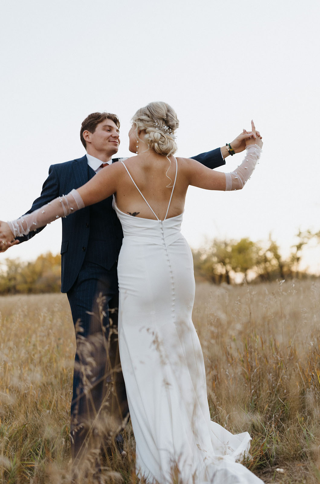 Back of bride in a fit and flare crepe wedding dress with buttons and sheer sleeves, dances with groom in a golden field.