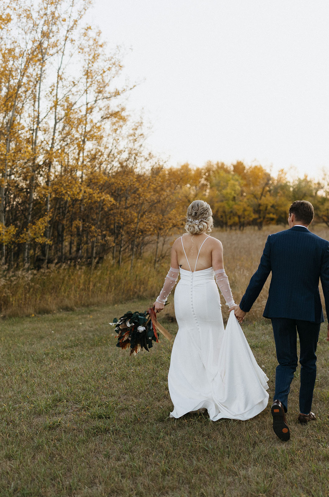 Bride in a fit and flare buttoned gown with thin straps and sheer sleeves, holds a bouquet and walks beside groom in a field.
