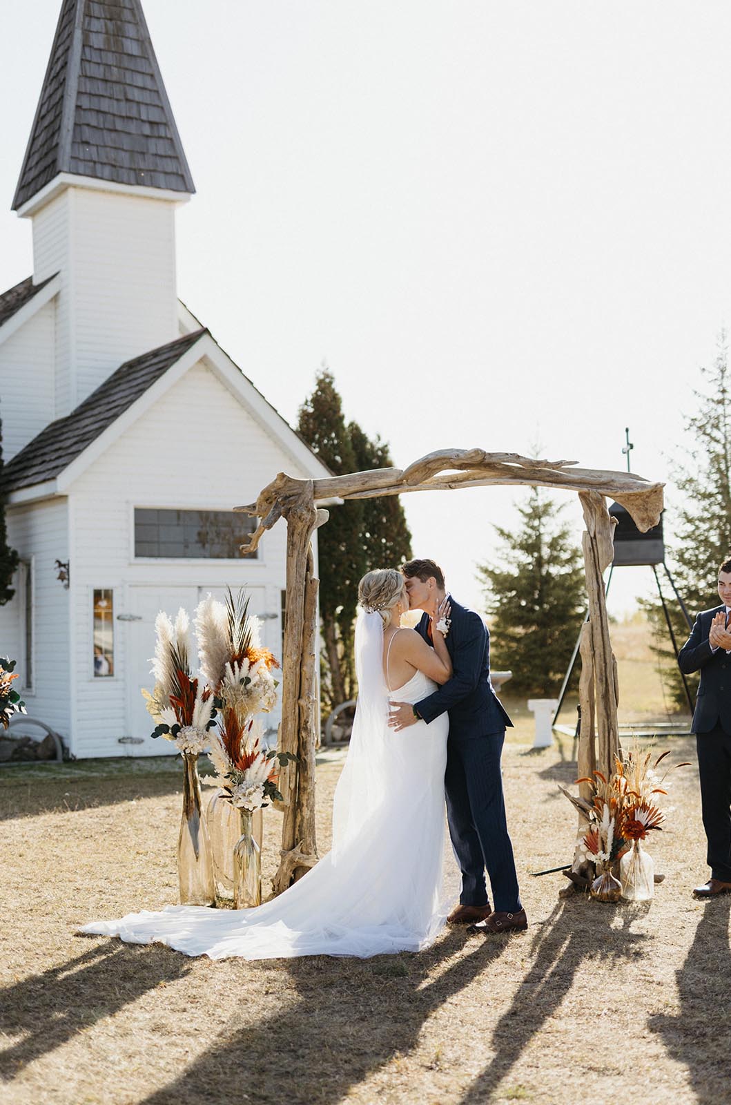 Groom kisses bride in a crepe spaghetti strap gown with fit and flare skirt and a veil, at a wooden altar, next to a chapel. 