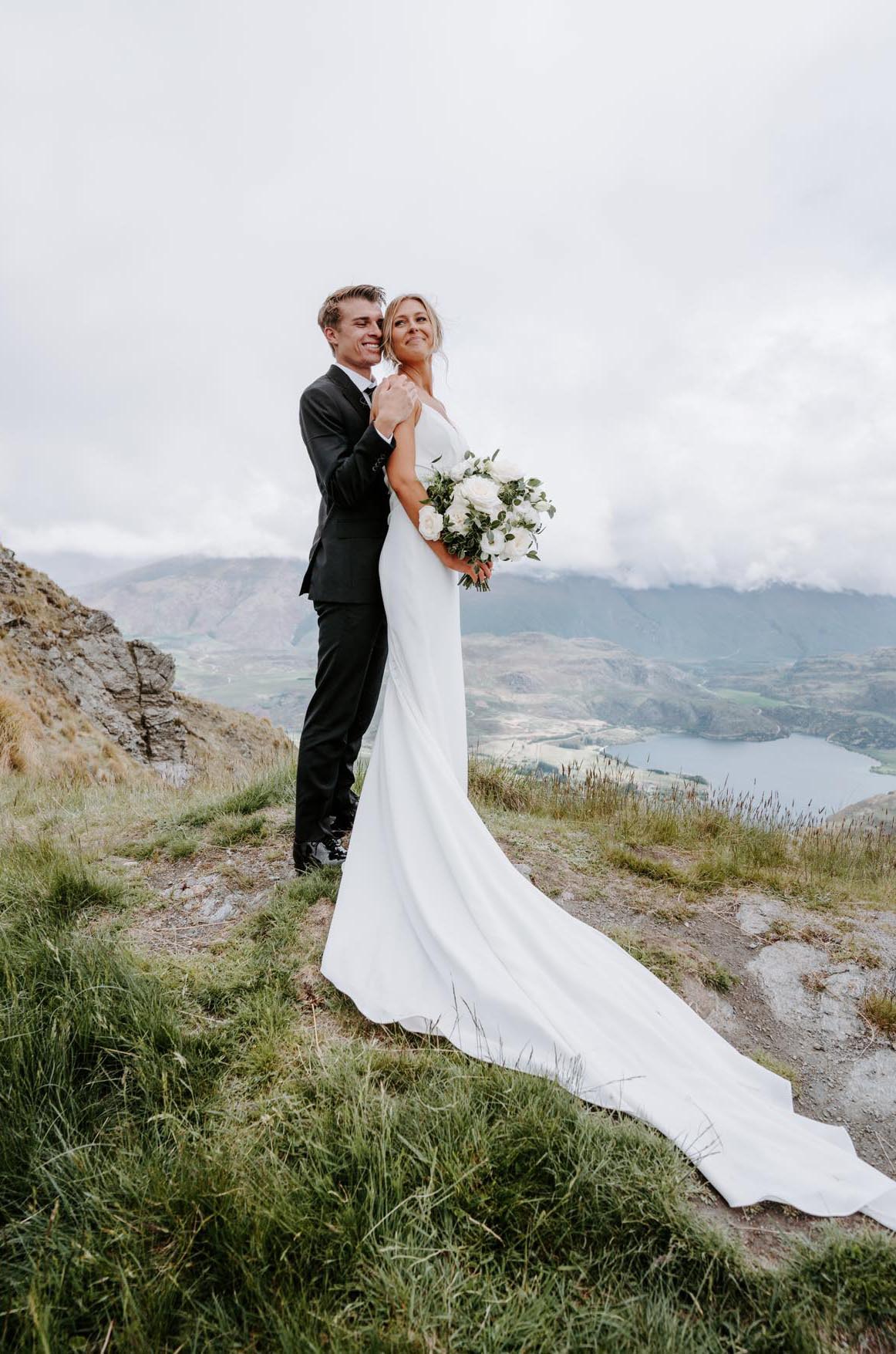 Groom embraces bride in a crepe fit and flare gown with a draped bodice, and is holding a bouquet on a mountain.