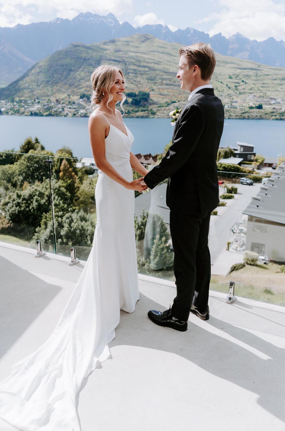 Bride in a crepe fit and flare wedding dress with thin straps, holds hands with groom on a terrace overlooking mountains.