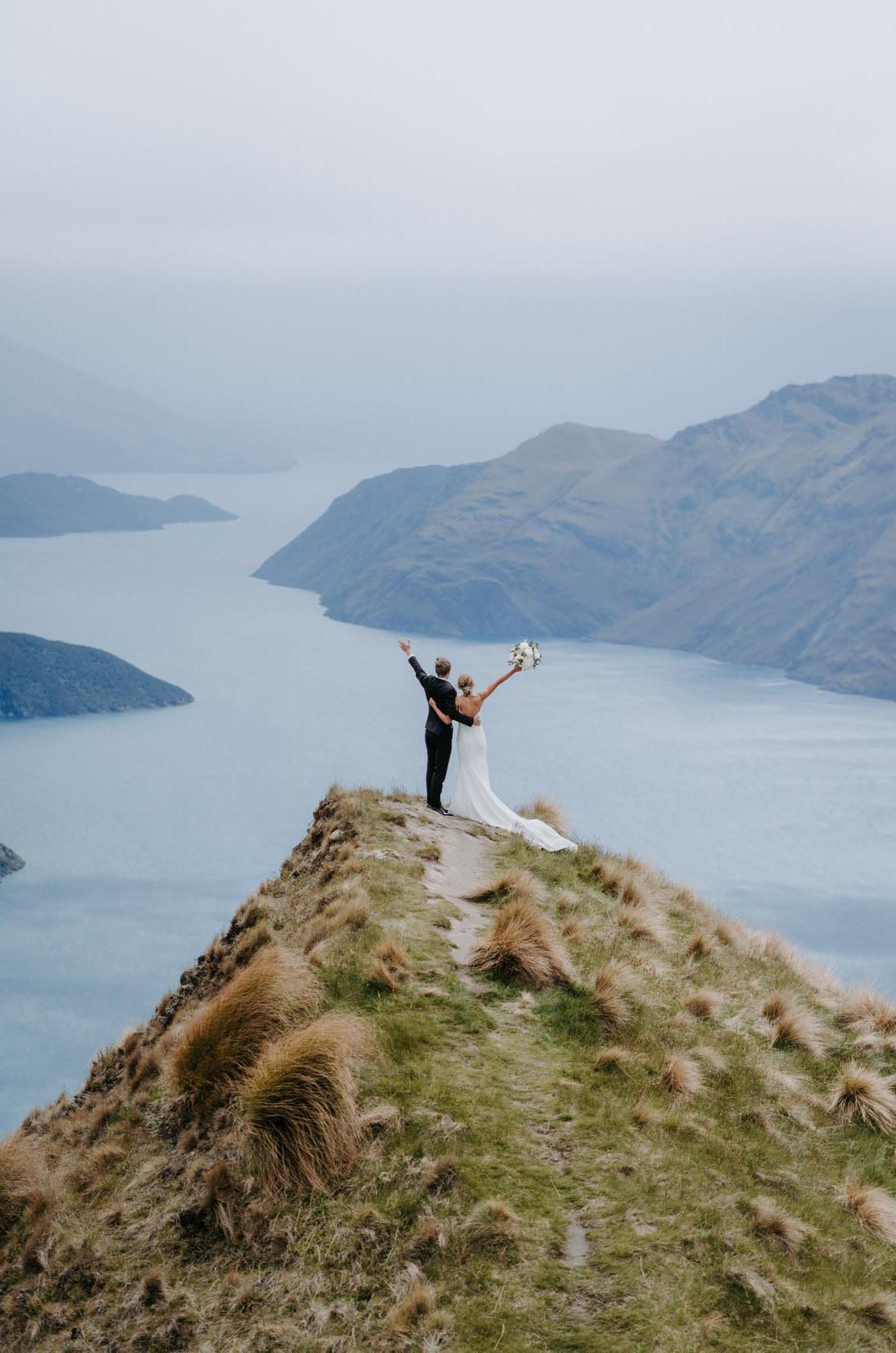 Bride in a crepe draped bodice gown with fit and flare skirt and a low open back, poses with groom on a mountaintop.