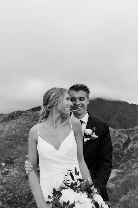 Close-up of bride in a draped bodice crepe gown with overlapping and thin straps, poses with groom in front of mountains.