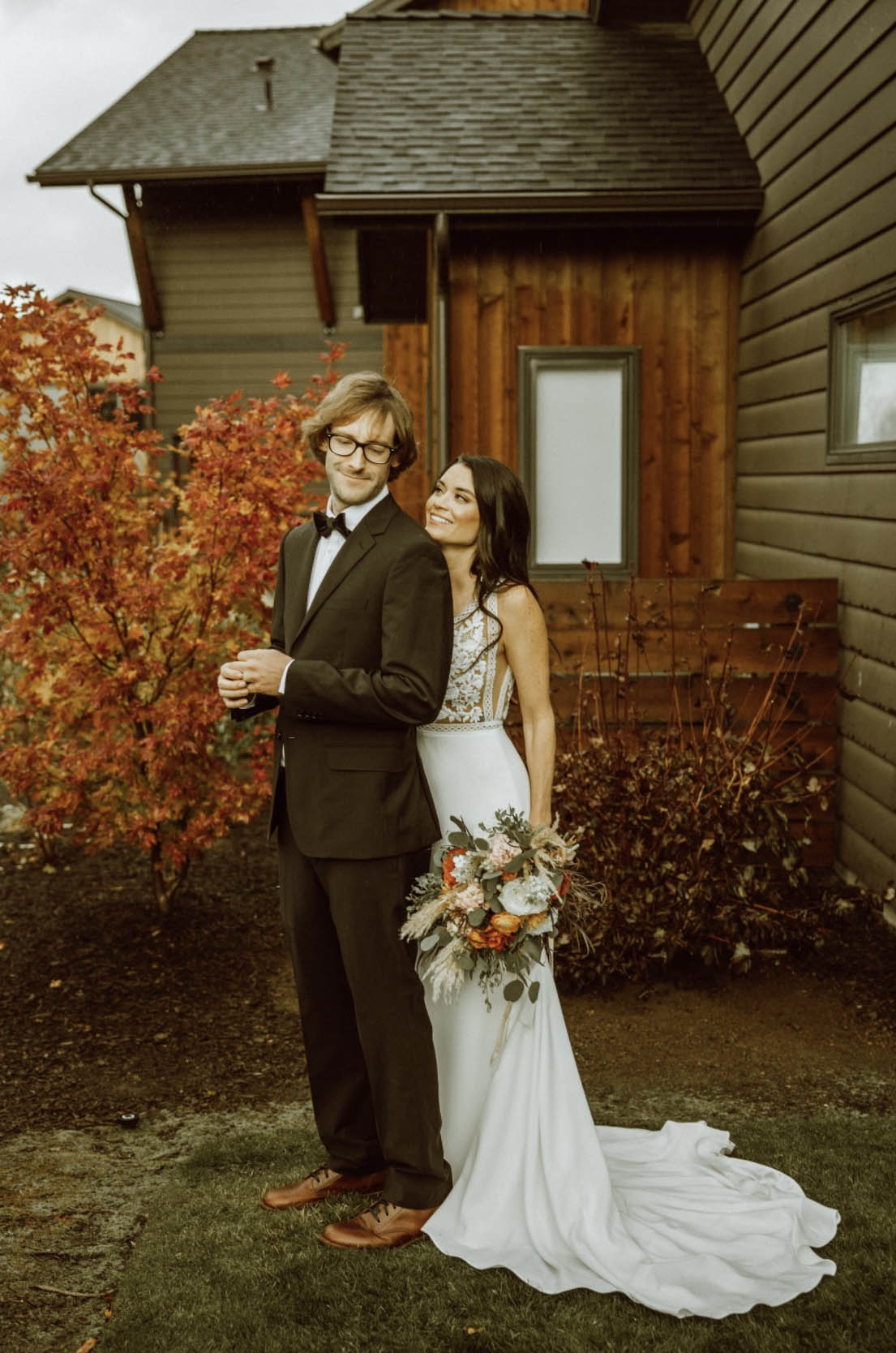 Bride poses in a lace and crepe gown with a belt and fit and flare skirt, holding a bouquet next to her groom in a tuxedo.