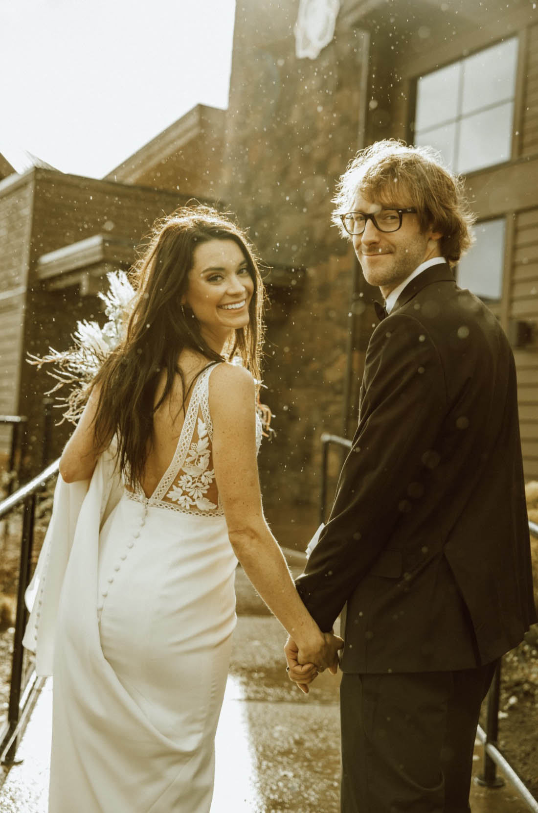 Back of bride smiling in a lace bodice wedding dress with a buttoned crepe skirt, walks alongside groom outside the venue.
