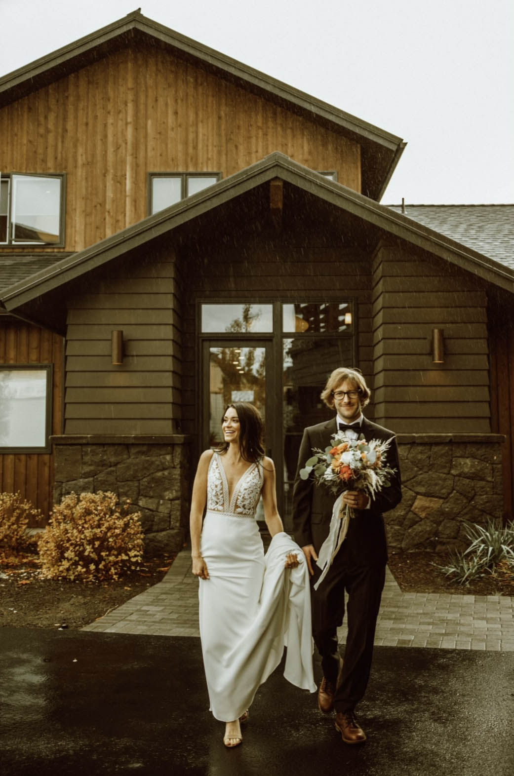 Bride in a lace bodice gown with a plunging neck and crepe skirt, holds her train and walks with groom in front of the venue.