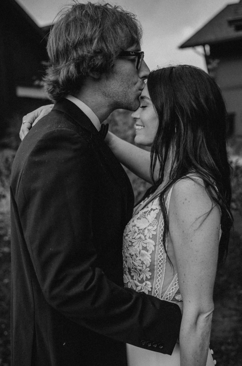 Close-up of groom embracing his bride smiling in a sleeveless lace gown with buttons and in black and white. 