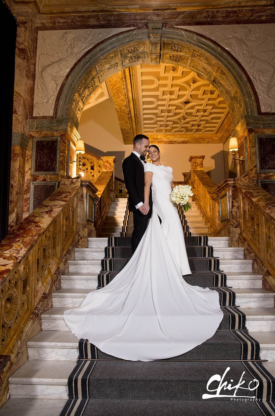 Bride in a bateau neck crepe gown with fit and flare skirt and flowing train, holds a bouquet beside groom on marble stairs.
