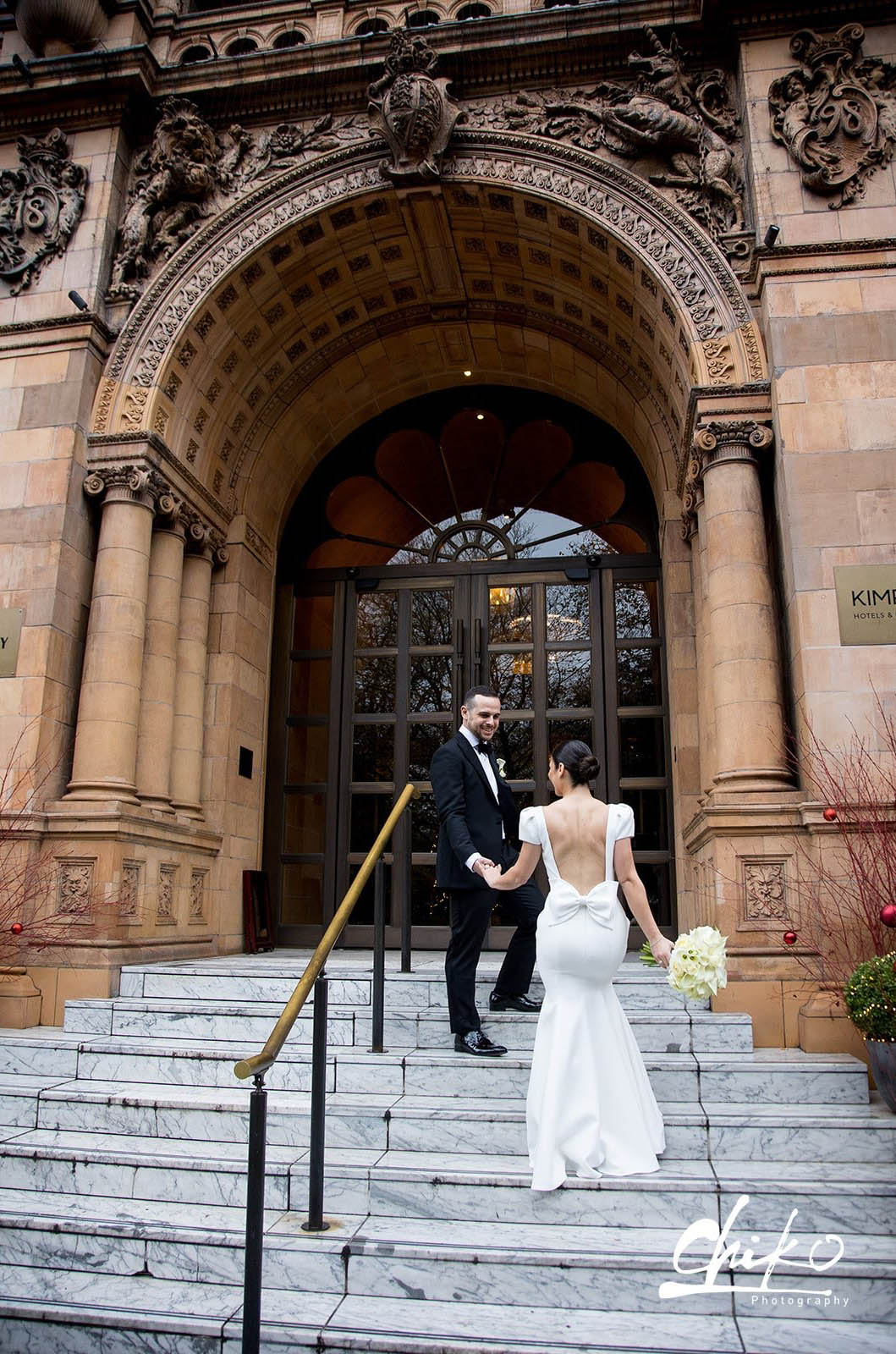 Bride in a low back crepe wedding dress with a bow and cap sleeves, holds a bouquet on a grand staircase with the groom. 