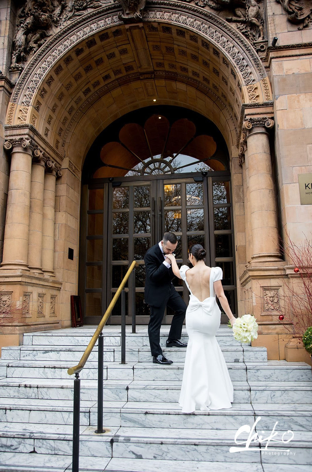Groom kisses bride's hand in a crepe fitted gown with a back bow, puff sleeves and an open back, on ornate marble stairs.