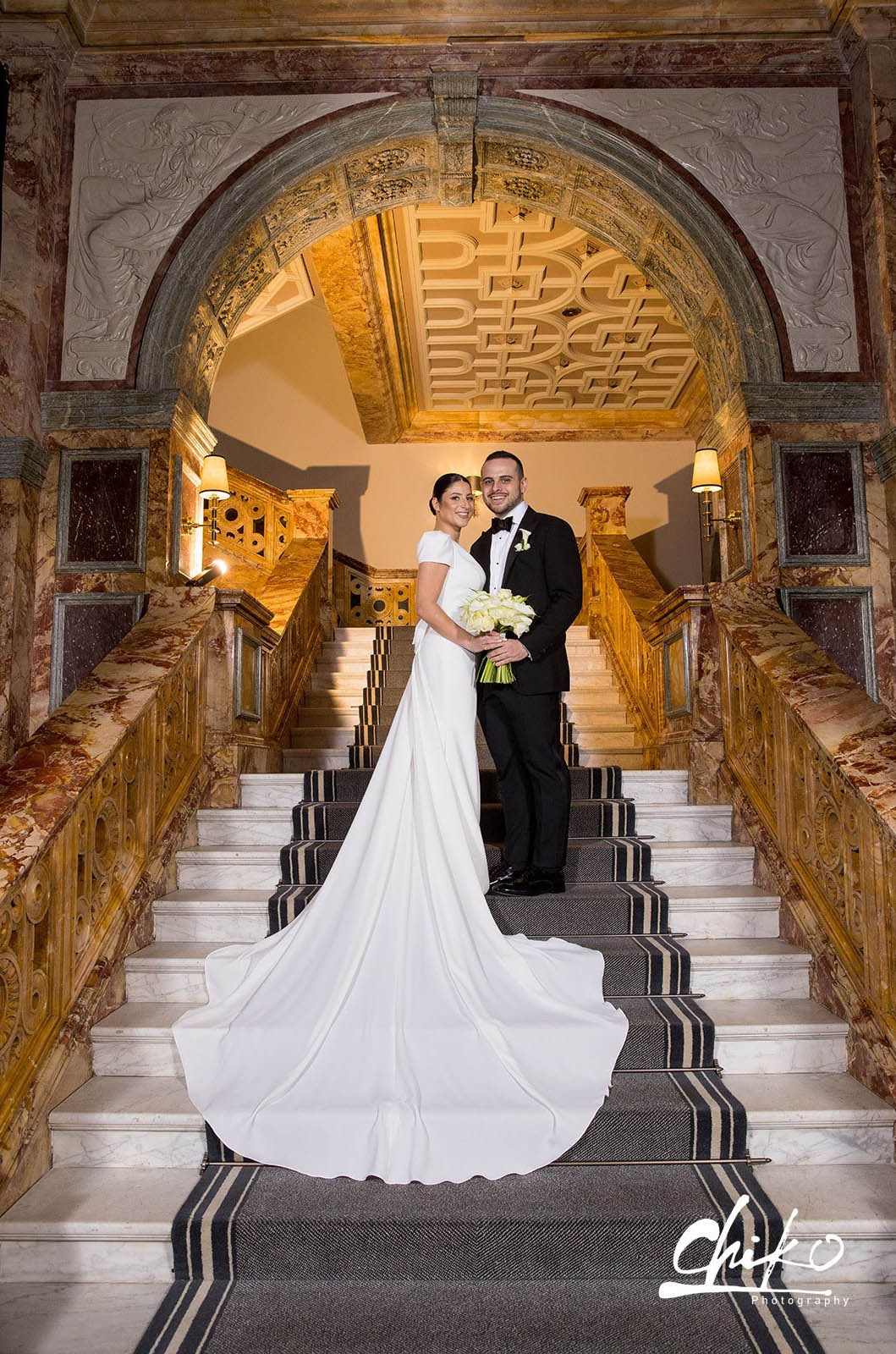 Bride in a crepe cap sleeve gown with a bow and fit and flare skirt, poses with groom on marble stairs under an ornate arch.