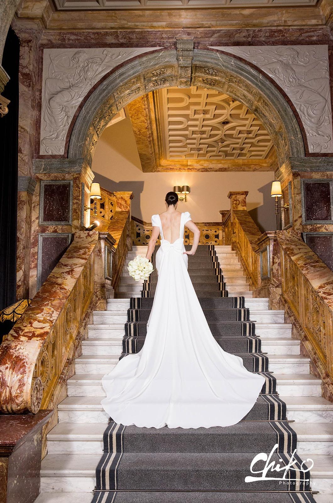 Bride poses in a crepe open back gown with a bow and flowing train, is holding a white bouquet on a grand winding staircase.