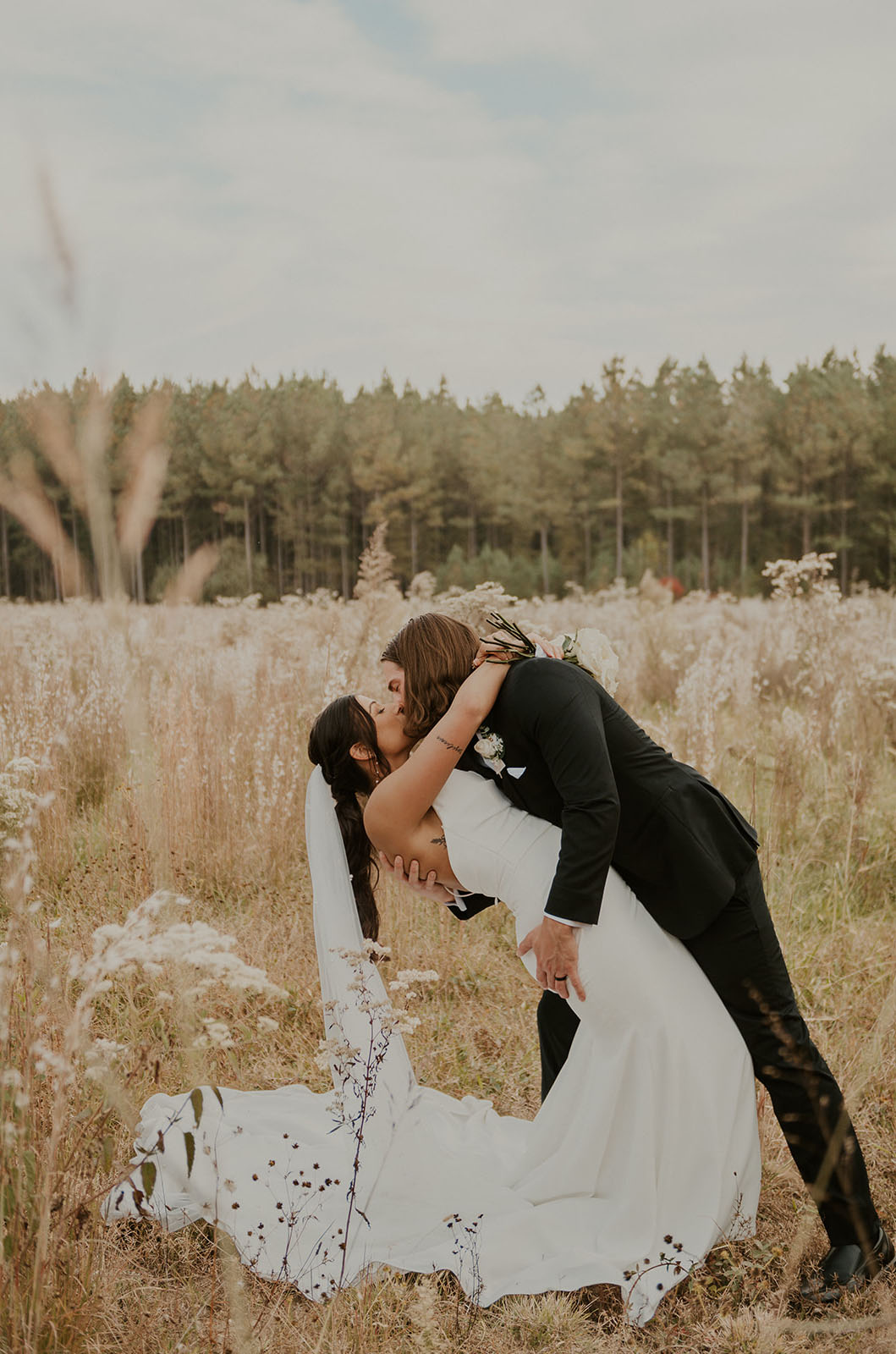 Groom dips his bride in a crepe fit and flare gown with a veil and a low back, in a golden field with trees.