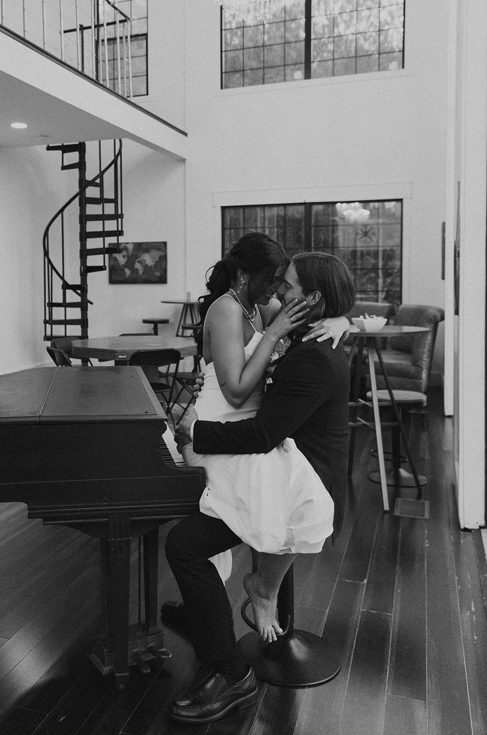 Bride in a spaghetti strap crepe wedding dress, shares intimate moment with groom sitting on a piano chair in a modern loft.
