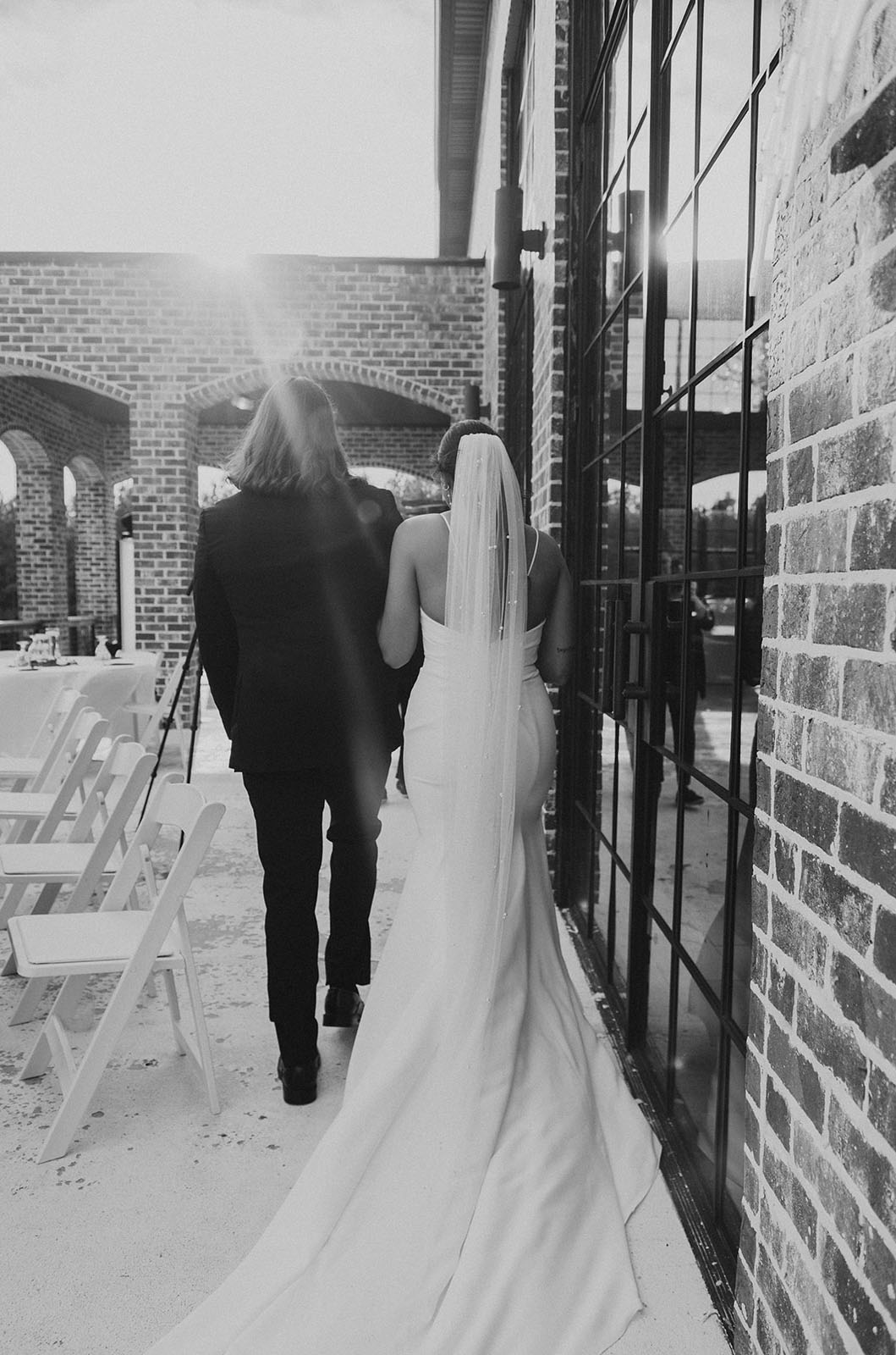 Bride in a sleeveless fit and flare gown with a long train and veil, walks alongside groom in black and white. 