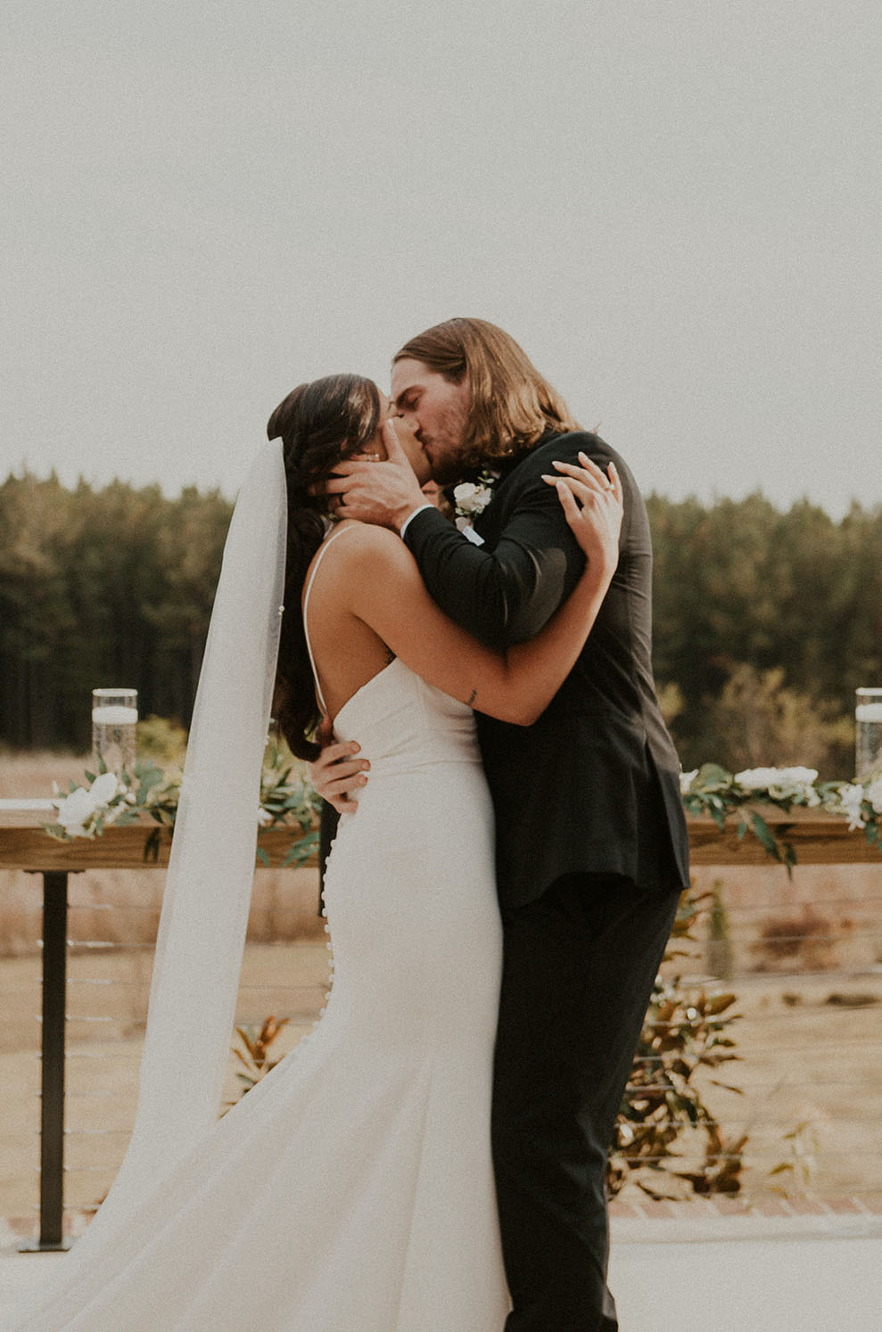 Groom kisses his bride in a crepe fit and flare wedding gown with thin straps, a veil and a low buttoned back outdoors.