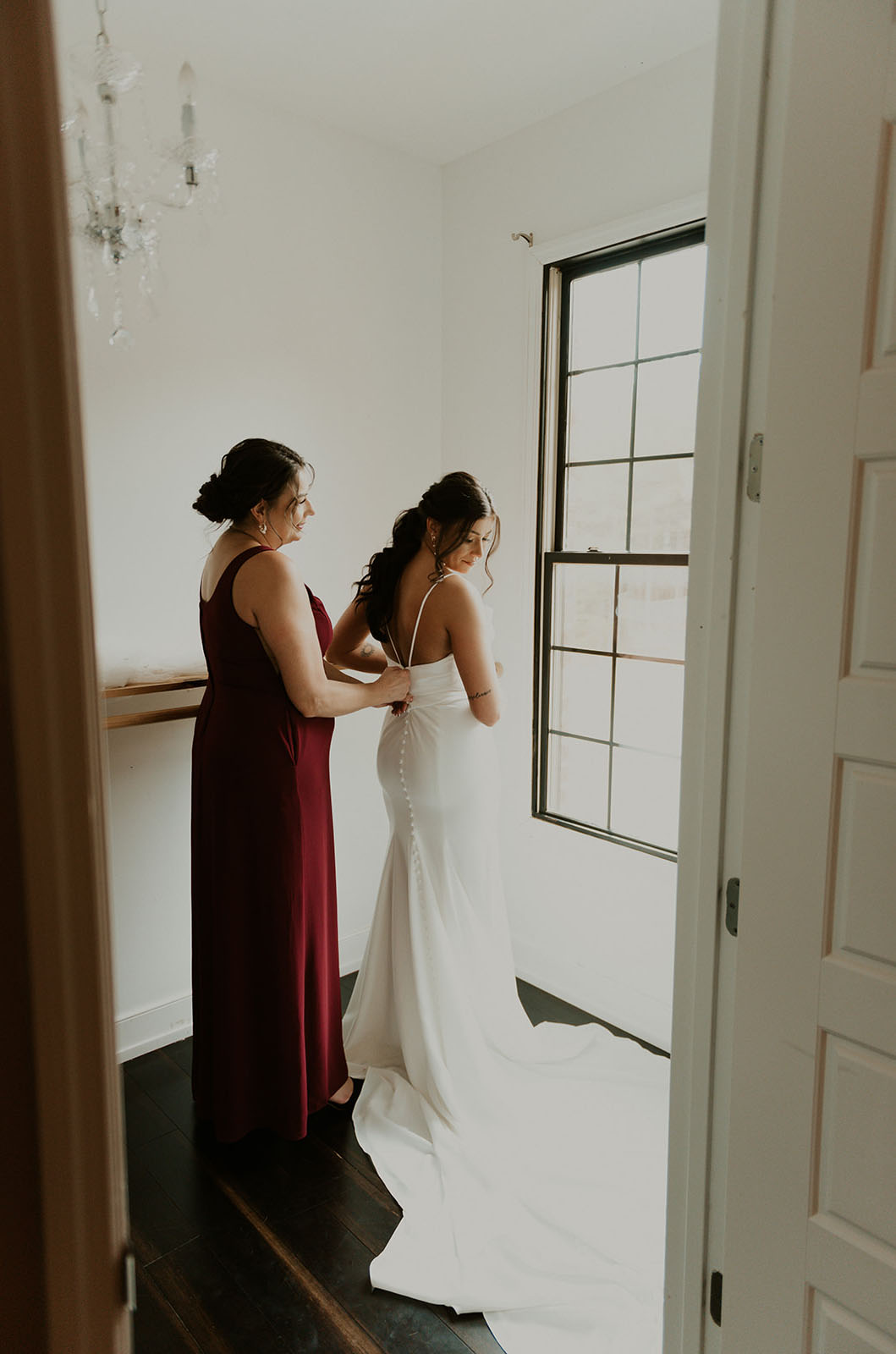 Bride is being fastened into a spaghetti strap crepe fitted gown with buttons and a flowing train, in a bridal suite.