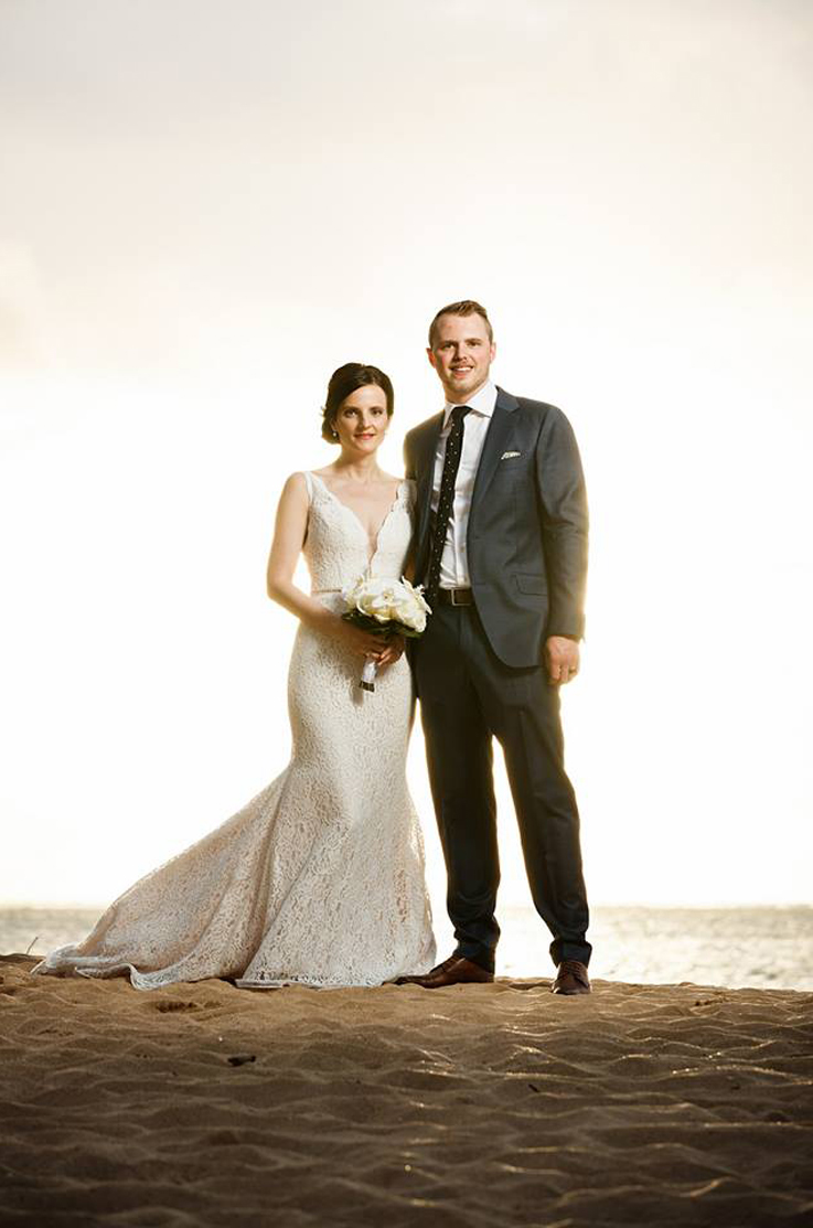 A bride in a plunging neckline lace wedding dress, holding a bouquet, and a groom in a suit stands on a sandy beach, with an ocean in the background.