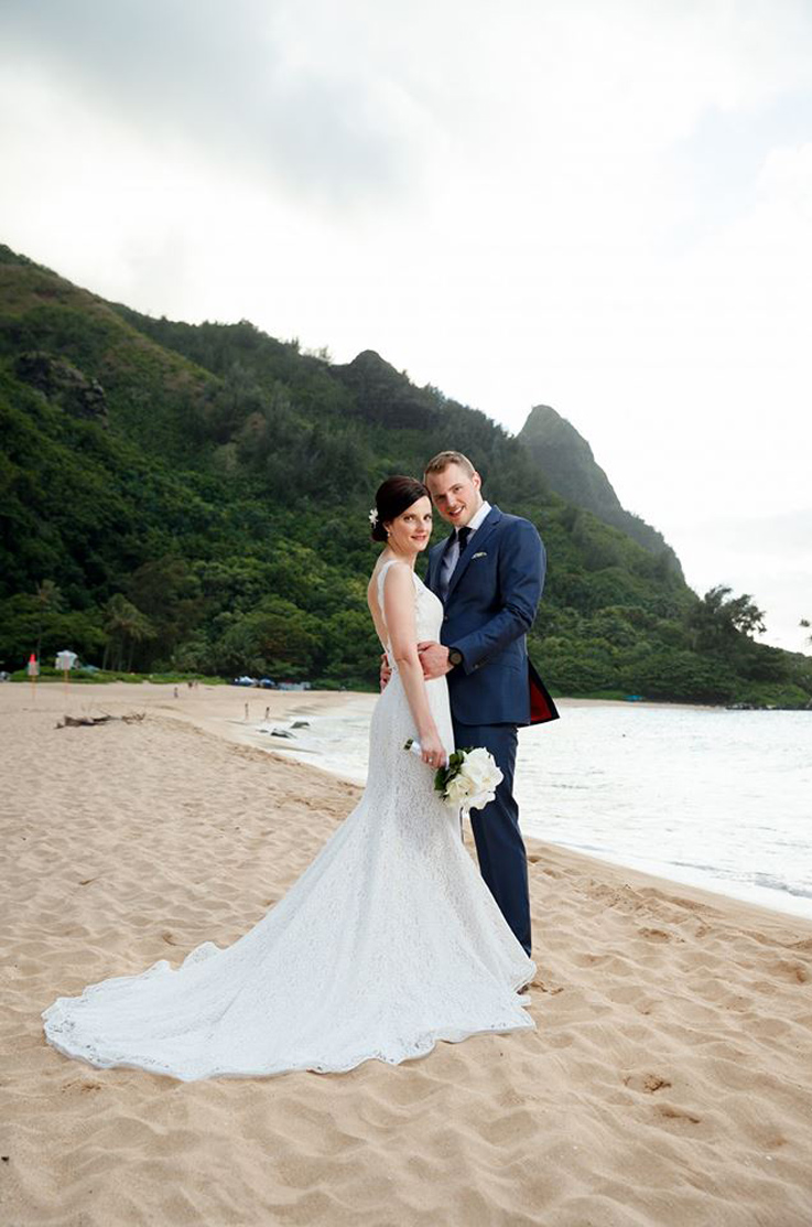 A groom in a suit holds the waist of a bride in a sleeveless lace wedding dress while they stand on the beach by the water, with tree covered hills in the background.