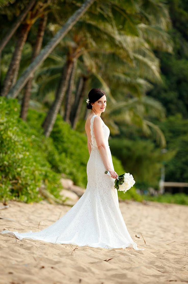 A bride in a sleeveless lace wedding dress stands on a sandy beach and looks over her shoulder while holding a bouquet.