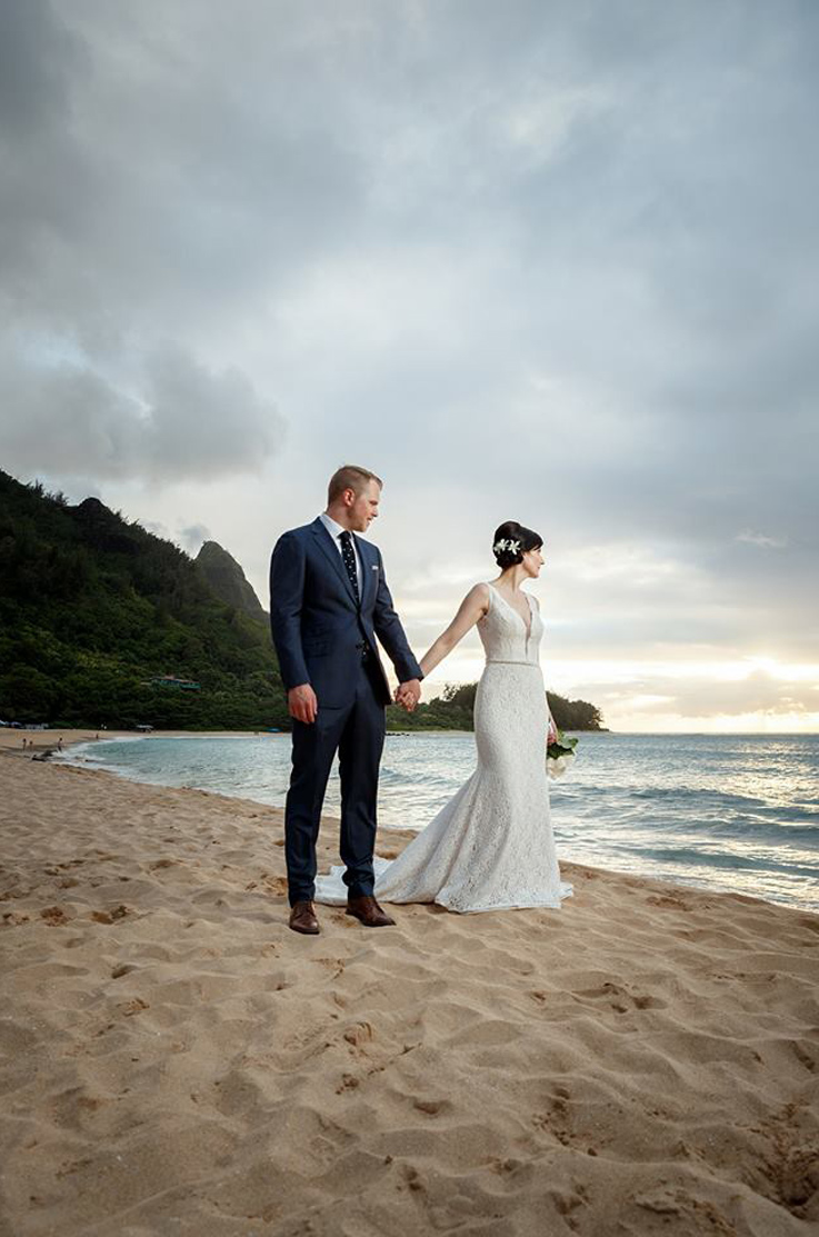 A bride in a plunging neckline lace wedding dress holds the hand of a groom in a suit as they stand on a sandy beach by the water, with trees and hills in the background. 
