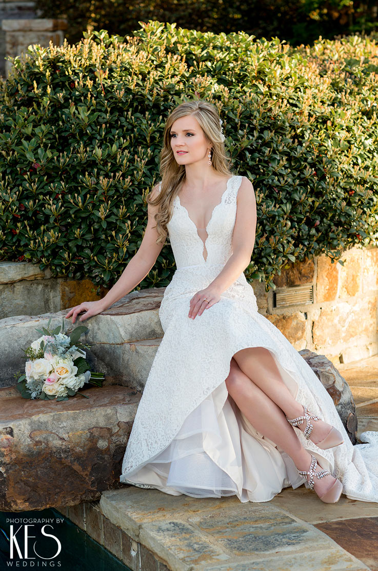 A bride in a sleeveless lace wedding dress its on stone steps and looks off to the side, with bushes behind her and her bouquet on the step beside her.