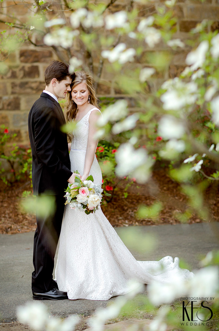 A bride in a sleeveless lace wedding dress, holding a bouquet, and a groom in a suit, stand on a path outdoors, with flowering tree branches in the foreground and flowers in the background.