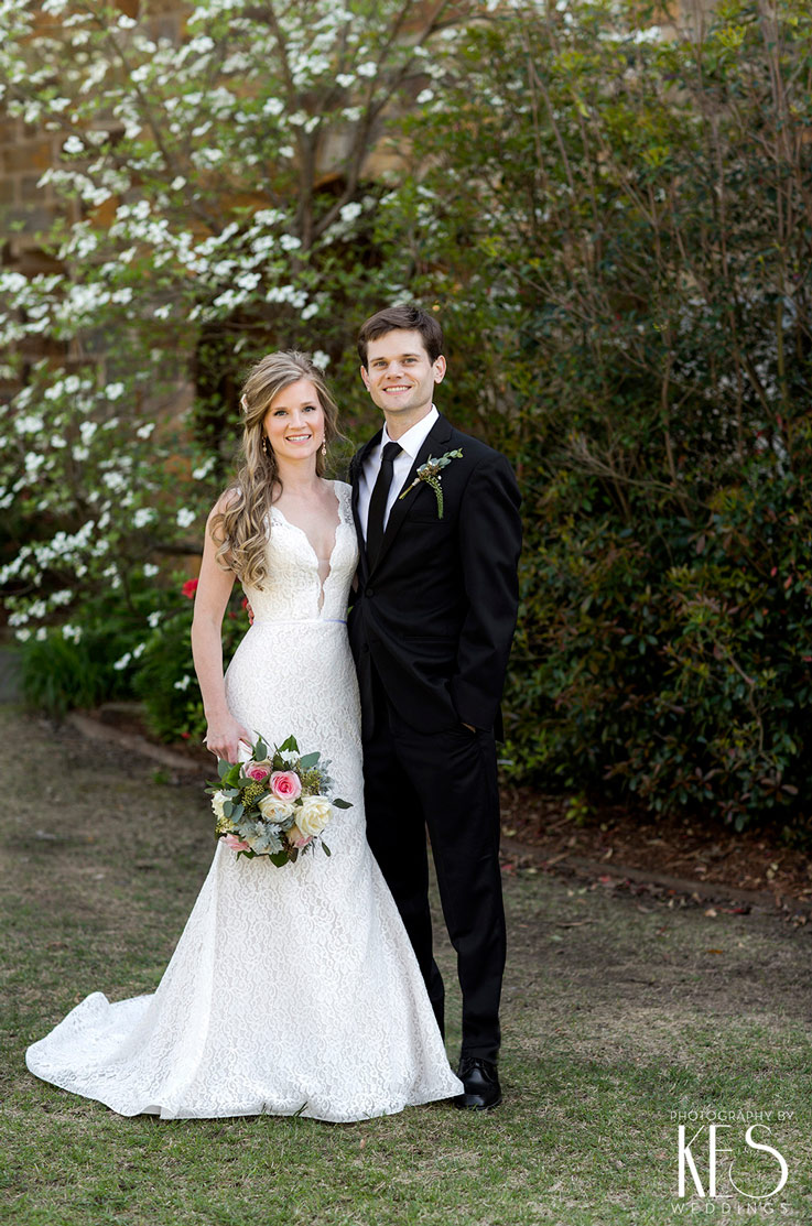 A bride in a plunging neckline lace wedding dress, holding a bouquet, and a groom in a suit stand together outside, with trees in the background.
