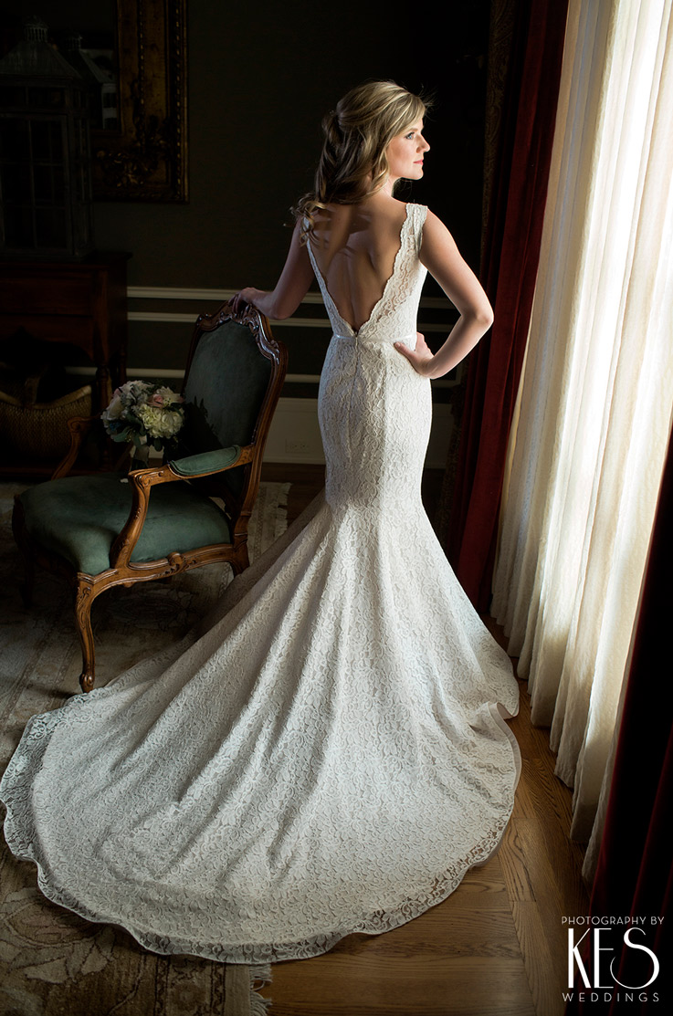 A bride in a sleeveless lace wedding dress stands indoors with her train fanned out behind her as she looks off to the side and leans against an ornate chair.