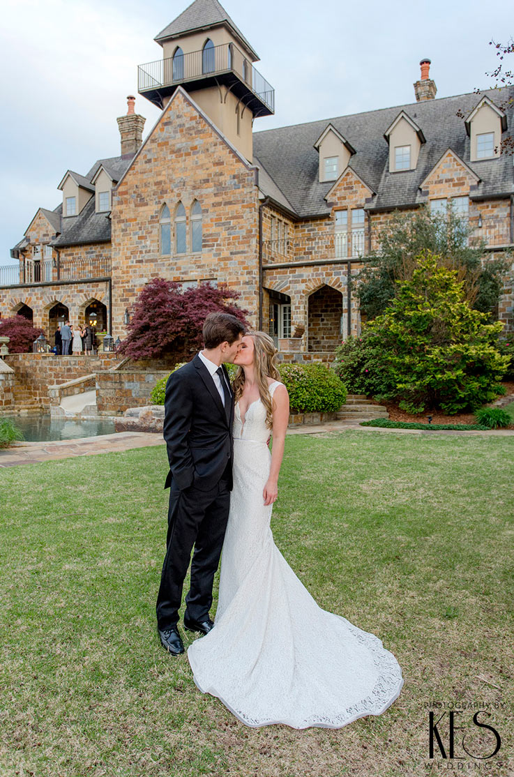 A bride in a plunging neckline lace wedding dress and a groom in a suit kiss outside, on the lawn of a brick mansion.