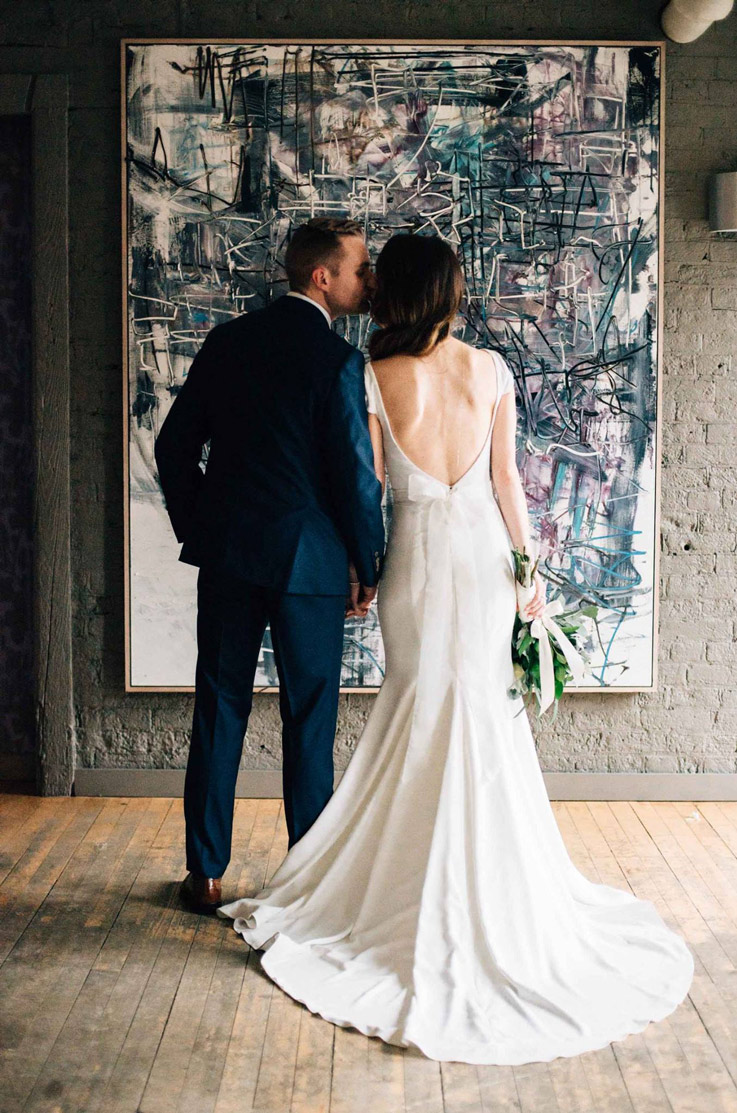 A bride in a cap sleeve wedding dress and a groom in a suit kiss while standing in front of abstract art.