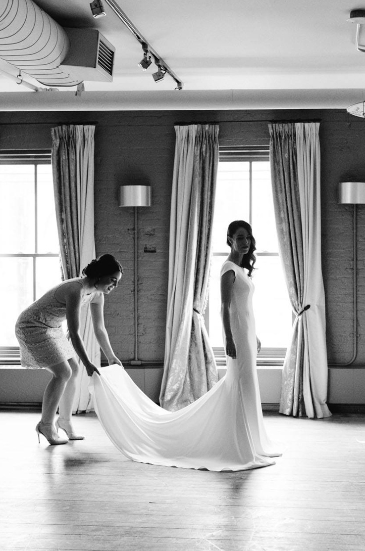 A woman in a lace dress fans out the dress train of a bride in a cap sleeve wedding dress, as they stand indoors in front of large windows.
