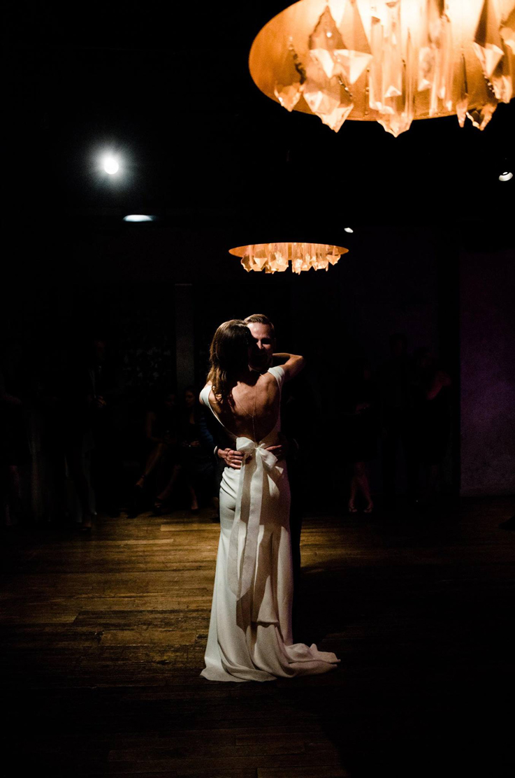 A bride in an open back wedding dress and a groom dances on a dimly lit dance floor, with wedding guests watching in the background.