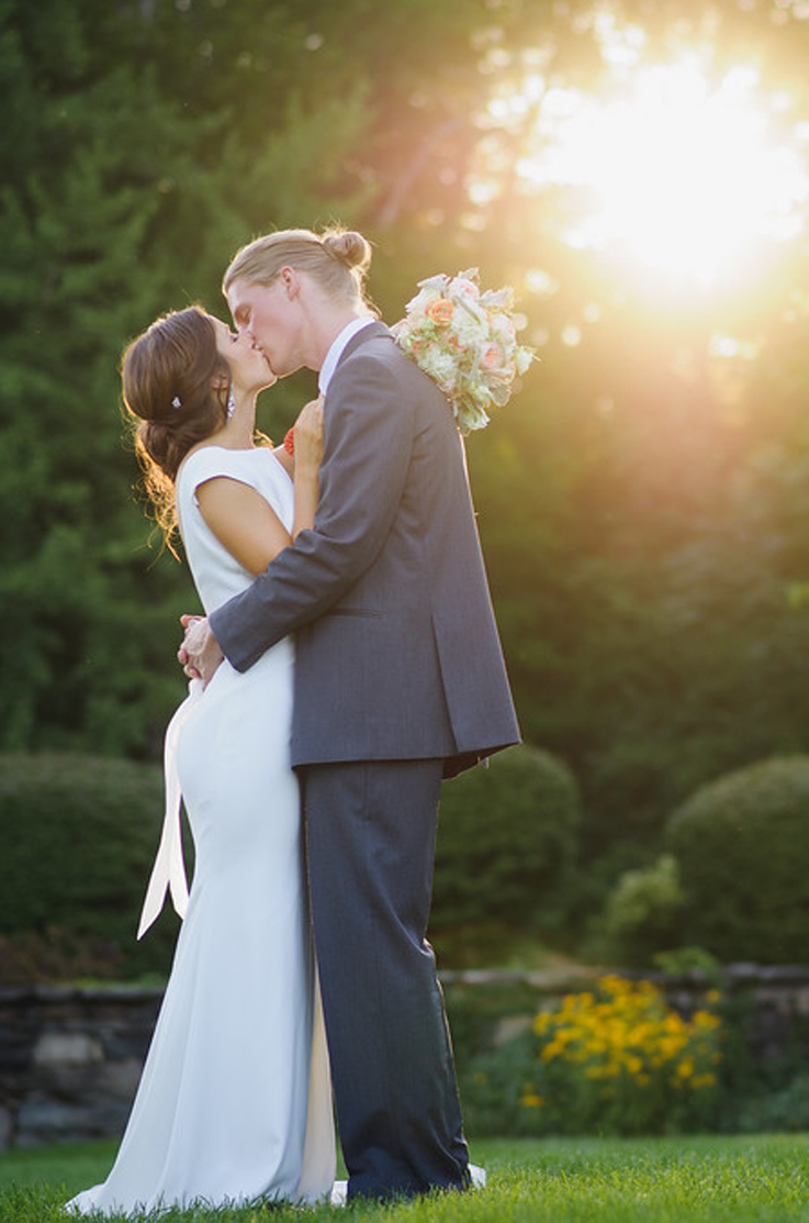 A bride in a cap sleeve wedding dress and a groom in a suit kiss outdoors with bushes, trees, and sunshine behind them.