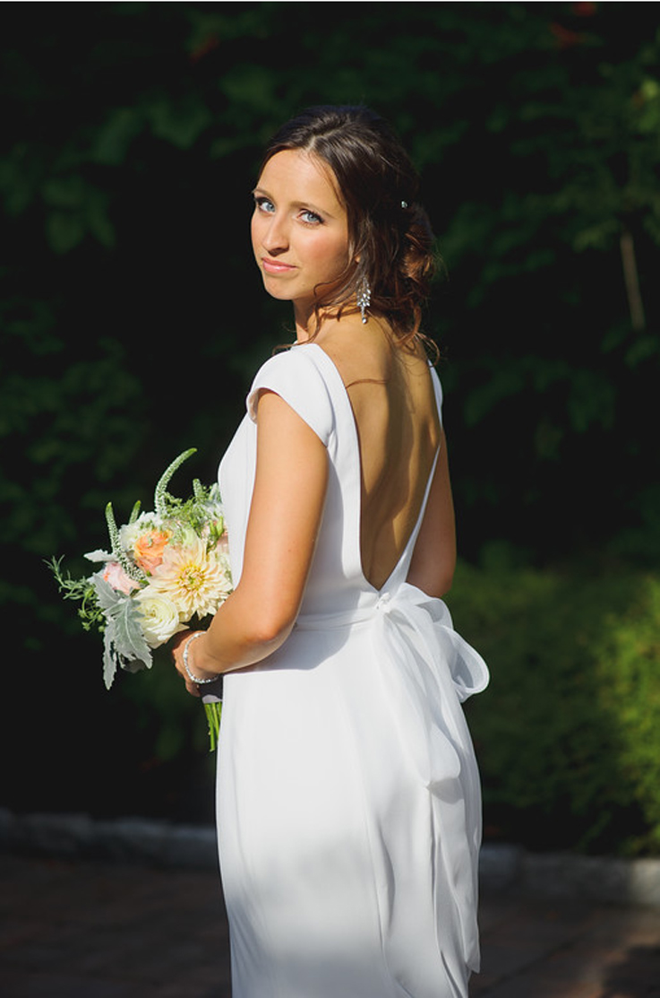 A bride in a cap sleeve wedding dress holds a bouquet and looks over her shoulder while standing outdoors in the sunlight.