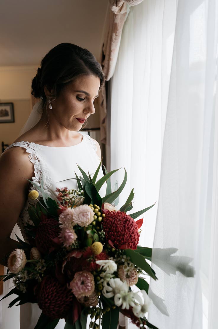 A bride in a sleeveless wedding dress holds a bouquet and looks down, while standing indoors in front of a window with sheer curtains.