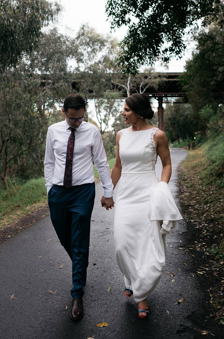 A bride in a sleeveless wedding dress and a groom in a dress shirt and tie walk down a path in a park outdoors, while holding hands and talking.