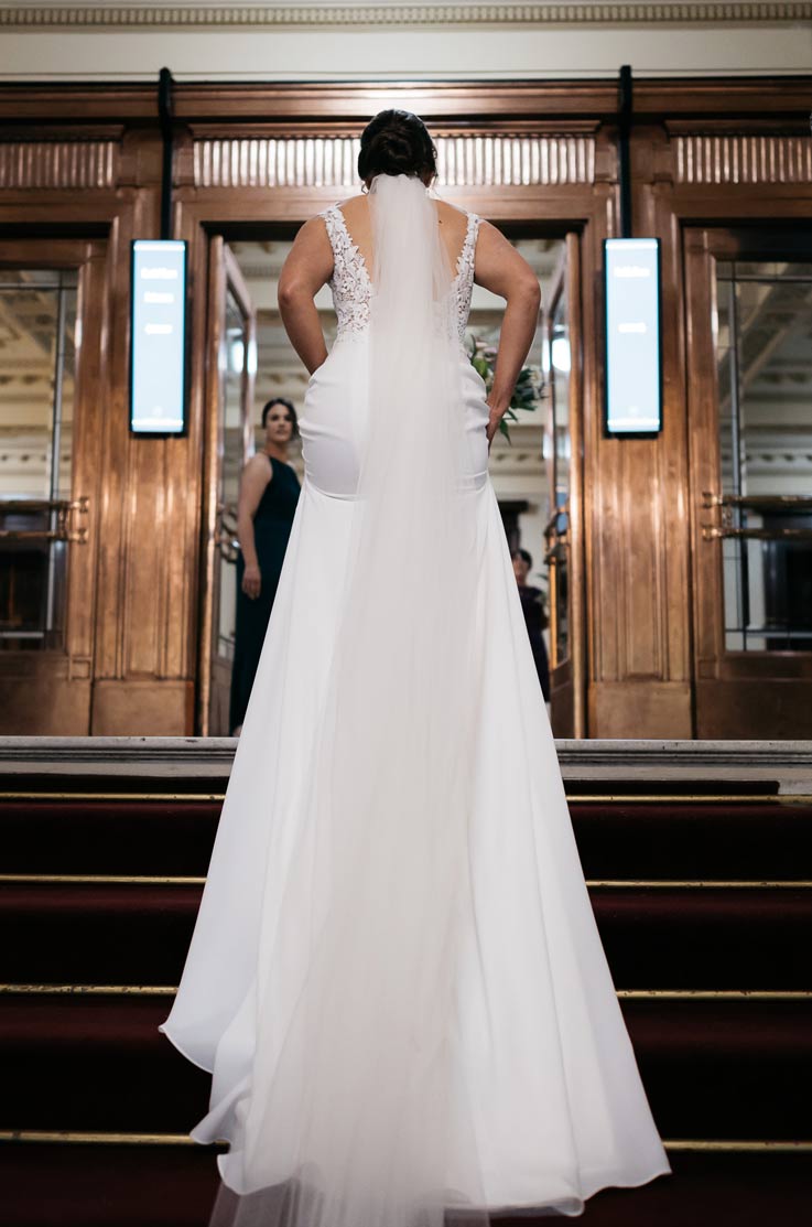 A bride in a sleeveless wedding dress and a veil walks up a stair case towards a multi-door entrance.