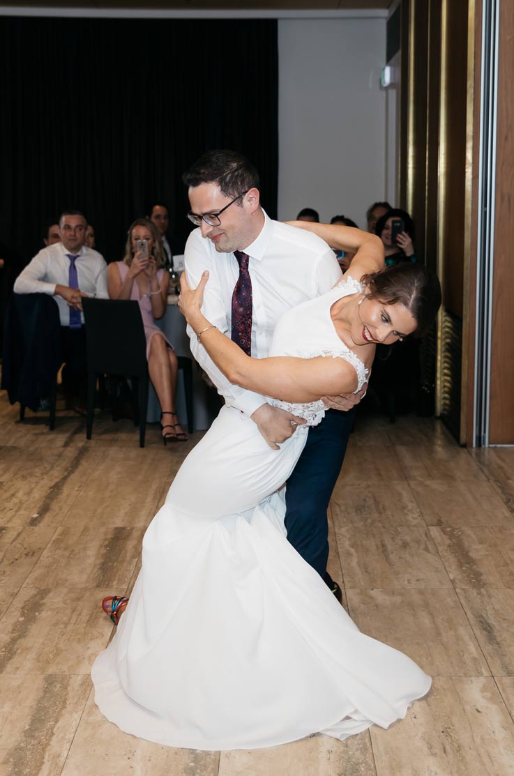 A groom in a dress shirt and tie dips a bride in a sleeveless wedding dress on an indoor dance floor, while wedding guests watch.