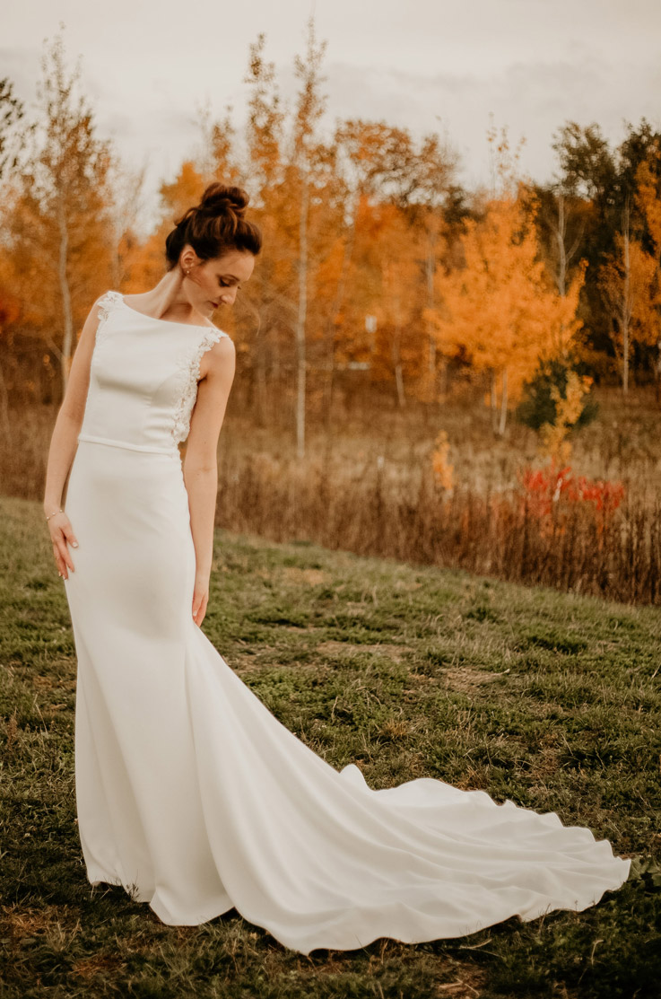 A bride in a sleeveless wedding dress poses outdoors in a grass field, her train fanned out behind her, with tall grass and trees behind her.