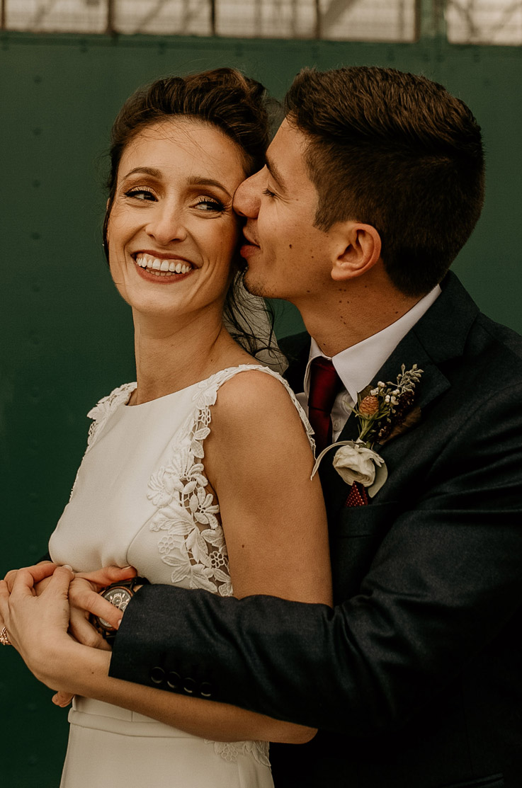 A groom in a suit hugs a bride in a sleeveless wedding dress from behind and kisses her on the cheek while she smiles.