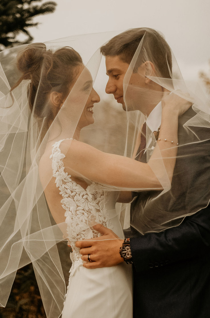 A bride in a sleeveless wedding dress and a groom in a suit look into each others eyes while holding each other, with the bride's veil over their heads.