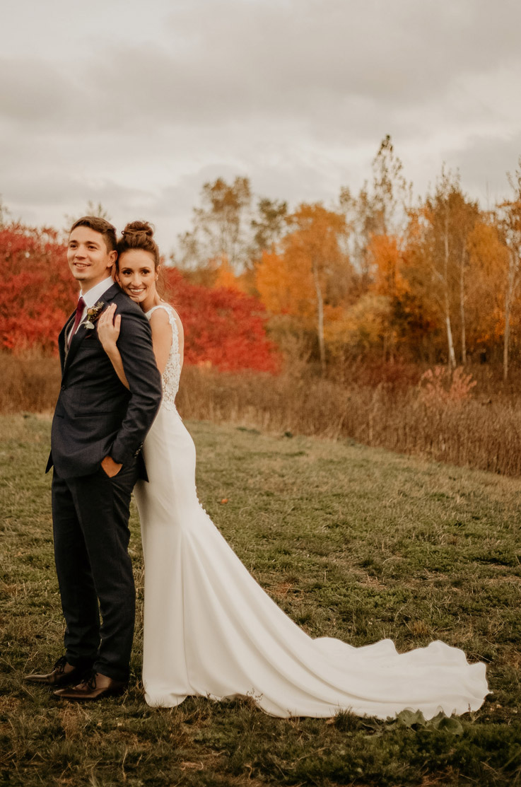 A bride in a sleeveless wedding dress holds a groom in a suit from behind as they both smile, while standing outdoors in a grass field with tall grass and trees behind them.
