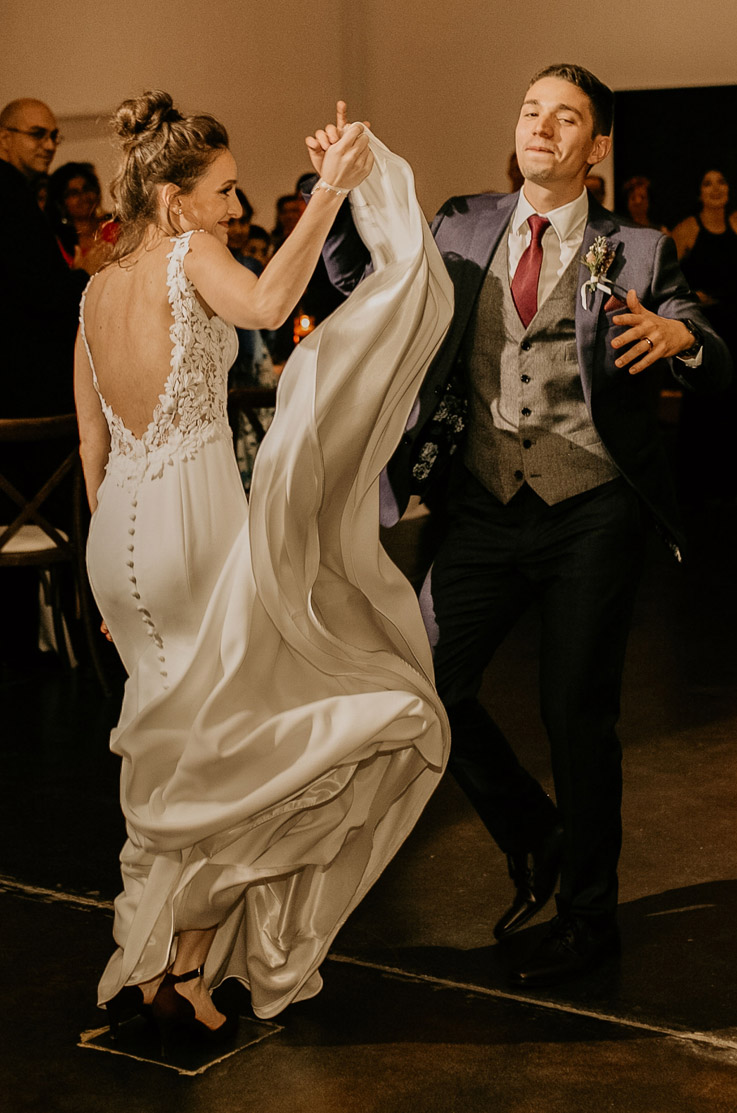 A bride in a sleeveless wedding dress holds the skirt of her dress with one hand while dancing with a groom in a suit on an indoor dance floor, with guests watching in the background.