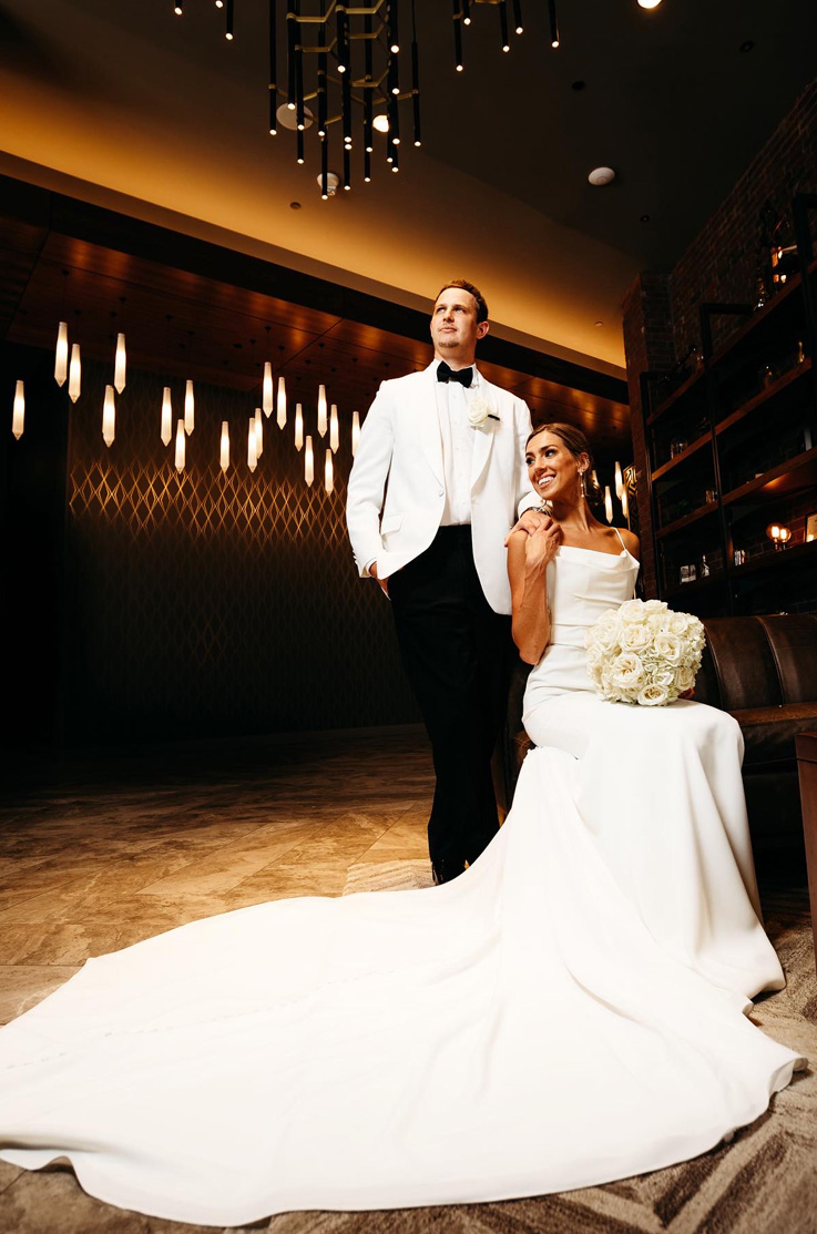 A bride in a long train wedding dress sits while a groom in a contrasting suit stands behind her, both are indoors in a dimly lit lounge.