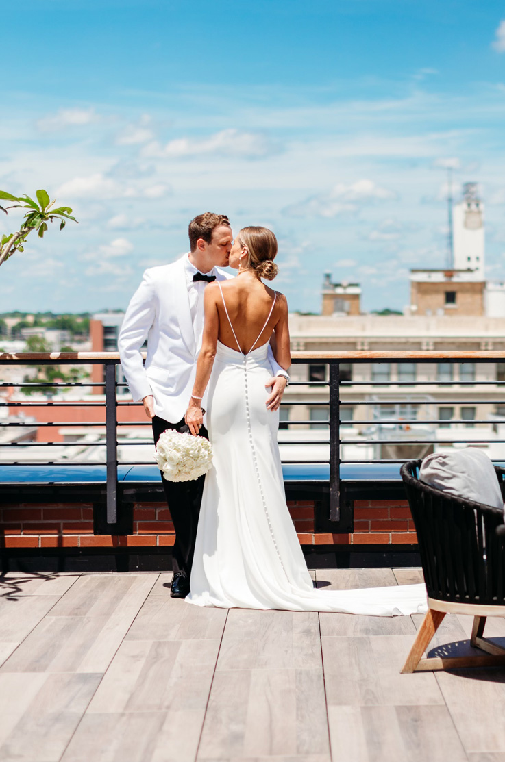 A bride in a long train wedding dress, holding a bouquet, and a groom in a contrasting suit kiss outdoors on a balcony, with a city in the background.