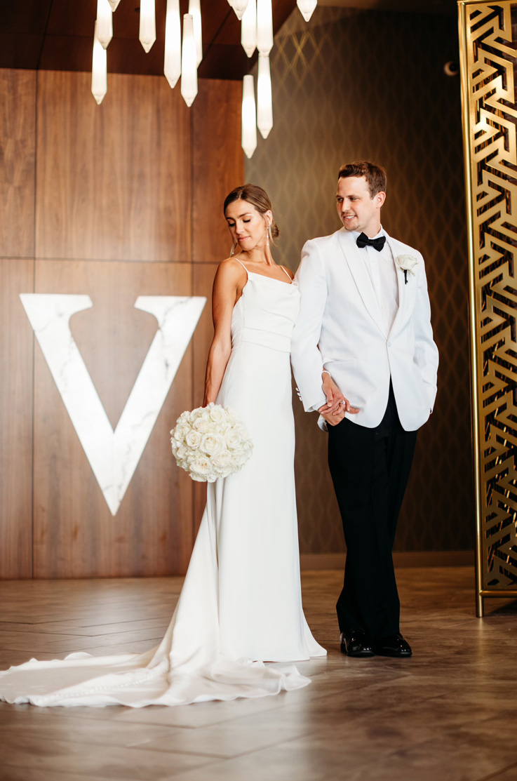 A bride in a long train wedding dress, holding a bouquet, and a groom in a contrasting suit hold hands and make an entrance. 