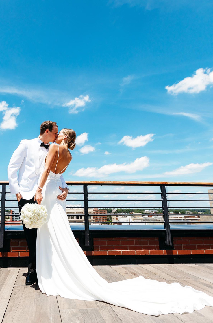 A bride in a long train wedding dress, holding a bouquet, and a groom in a contrasting suit kiss outdoors on a balcony, with a city in the background.