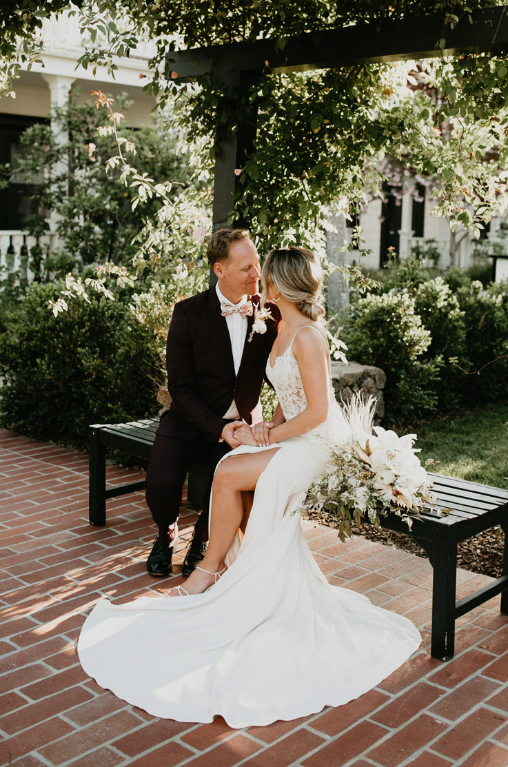 A bride in a skirt slit wedding dress and a groom in a suit sit on a bench outdoors and hold hands while looking into each others eyes.